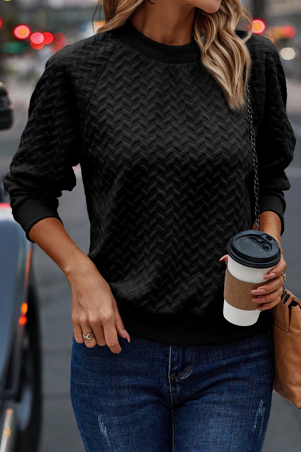 A person with long hair, wearing a Black Solid Textured Raglan Sleeve Pullover Sweatshirt and blue jeans, is holding a beige clutch while standing on a city street, showcasing their versatile style.