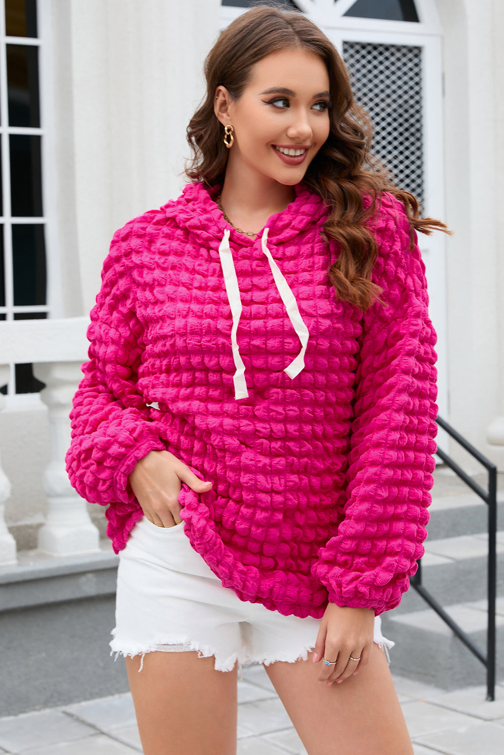 A person stands with their back to the camera, donning the Rose Bubble Textured Waffle Hoodie in a vibrant pink hue. They are in a room with yellow floral wallpaper, and a corner of a wooden chair is visible.