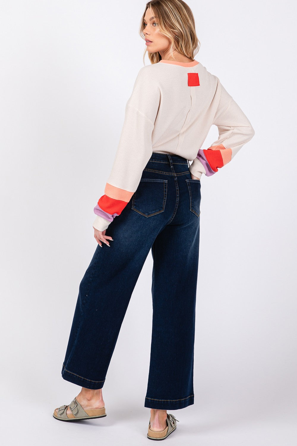 A person wearing a beige sweater, SAGE + FIG High Waist Wide Leg Jeans, a gray hat, and sandals is standing against a plain background.