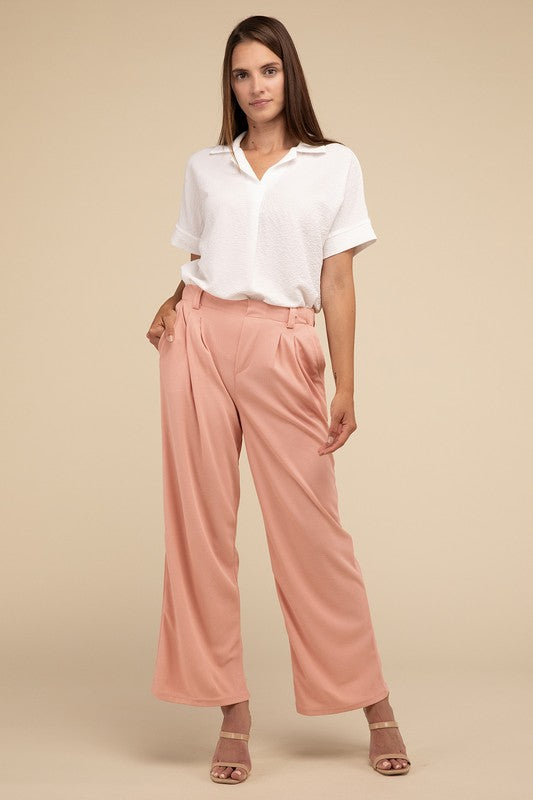 A woman stands against a beige background, highlighting her chic ensemble with a tailored fit. She wears a white short-sleeve shirt paired with versatile pink Waffle Trouser Pants, hands casually in pockets, complemented by stylish strappy sandals.