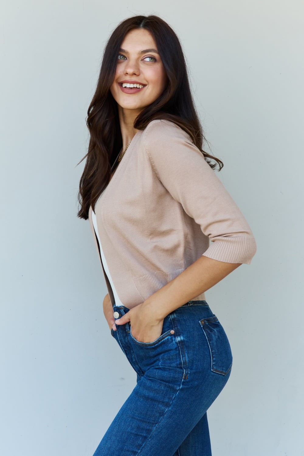 Woman with long dark hair wearing the versatile Doublju My Favorite Full Size 3/4 Sleeve Cropped Cardigan in Khaki over a white top and blue jeans, standing against a plain background.