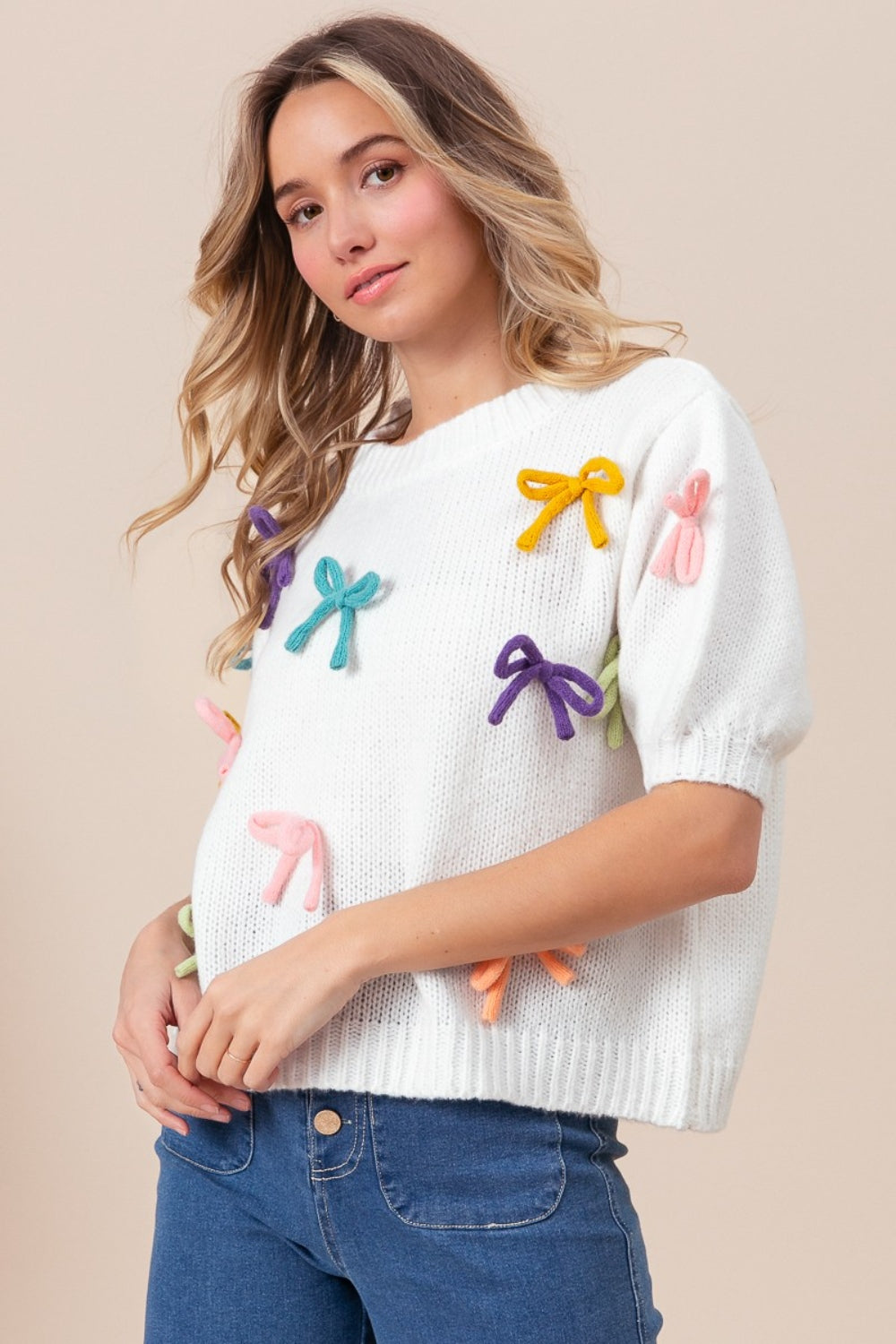 A woman stands against a beige background, wearing the BiBi Bow Detail Puff Sleeve Sweater in cozy white fabric with colorful bow decorations, paired with blue jeans.