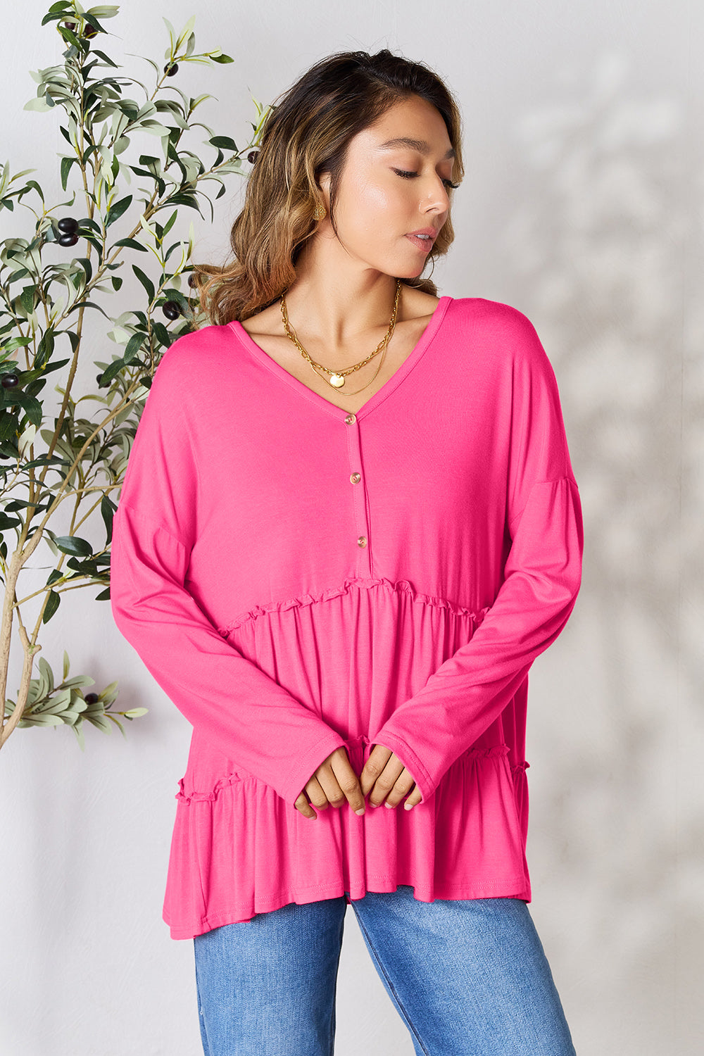 Person stands indoors near a plant, wearing the Double Take Half Button Long Sleeve Ruffle Hem Blouse in gray and blue jeans, smiling while looking to the side. The slightly stretchy fabric of the blouse adds comfort while its buttoned design offers a touch of elegance.