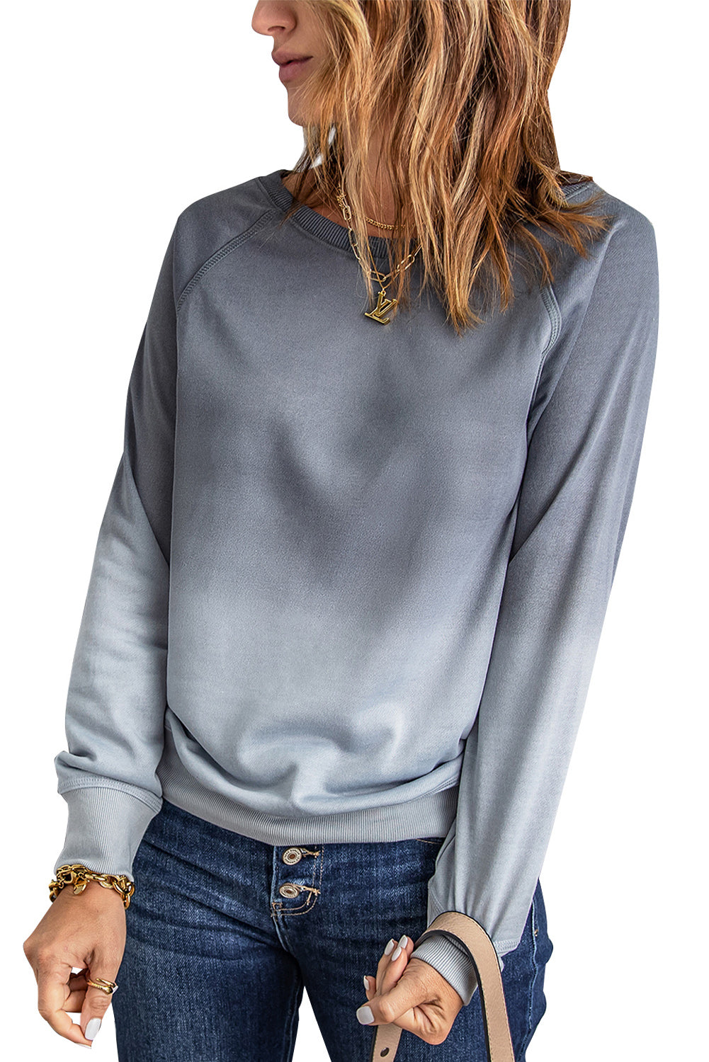 A person with wavy hair stands facing a gray wall, wearing a Gray Ombre Crewneck Long Sleeve Sweatshirt and blue jeans, with one hand on their hip.