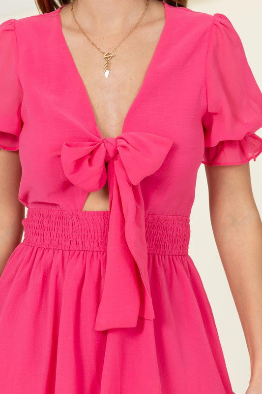 A woman is wearing the "Days Like This Tie-Front Mini Dress" in a vibrant pink color, featuring short ruffled sleeves, a prominent bow at the chest, and a tiered skirt with a smocked waist.