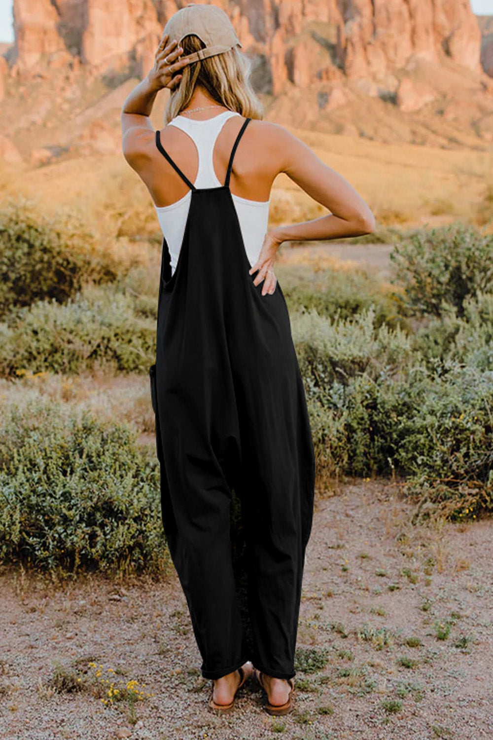 Person wearing a Double Take Full Size V-Neck Sleeveless Jumpsuit with Pockets and sandals, walking outdoors on a dirt trail with bushes in the background.