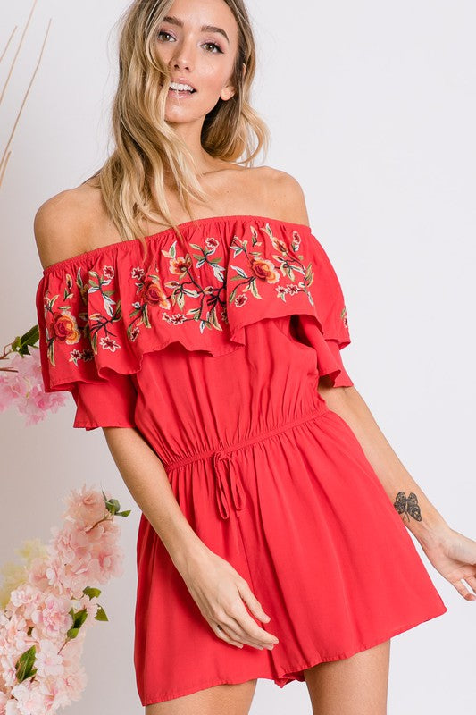 Wearing the stylish Floral Embroidered Off Shoulder Romper, a woman poses gracefully, showcasing its ruffle detail and making it an ideal addition to any summer wardrobe.