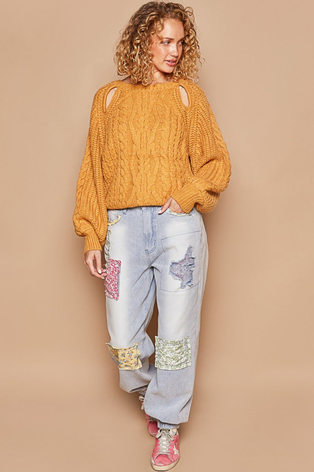 A person with curly hair wears a POL Cable Knit Cutout Long Sleeve Sweater in mustard, paired with light blue jeans adorned with patchwork designs.