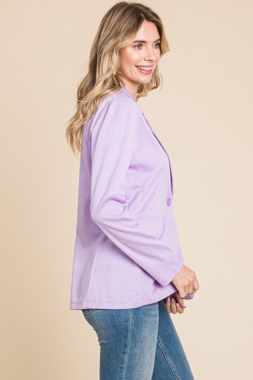 A person with long, wavy hair is wearing the Culture Code Button Up Long Sleeve Slit Blazer in light purple over a white top and blue jeans, standing against a beige background.