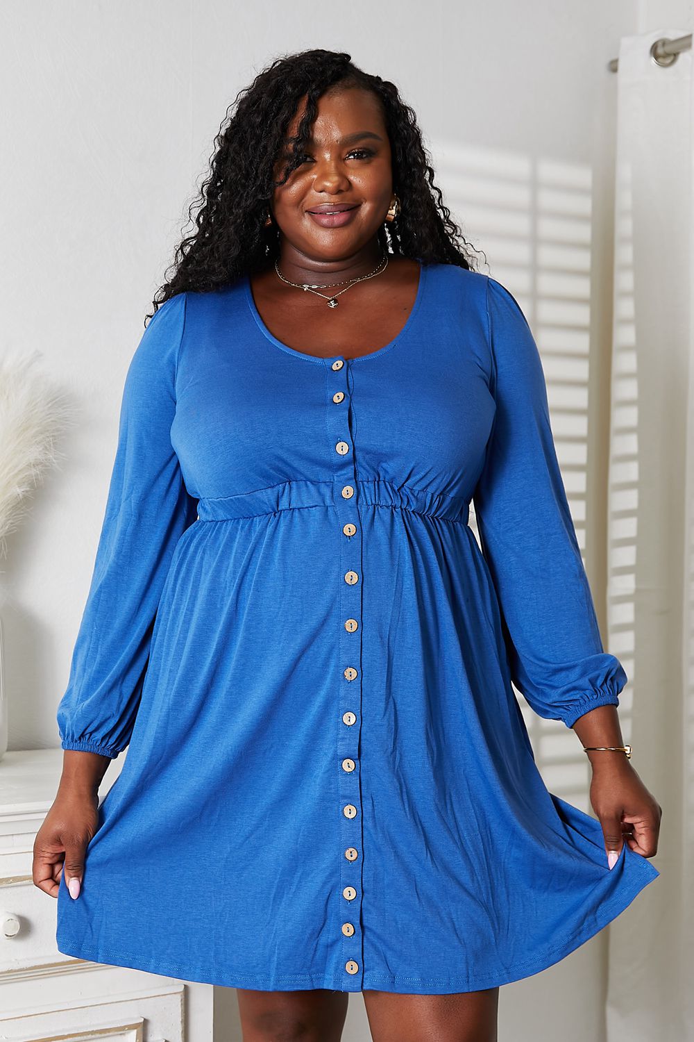 A woman poses indoors, smiling and holding out the skirt of a blue Double Take Scoop Neck Empire Waist Long Sleeve Magic Dress. She has curly hair and stands in front of a white wall with a partially visible piece of furniture.