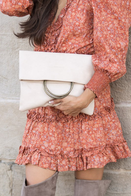 A person holds a tan Fold-over O-ring Clutch with a circular handle in front of their floral skirt and cream top.