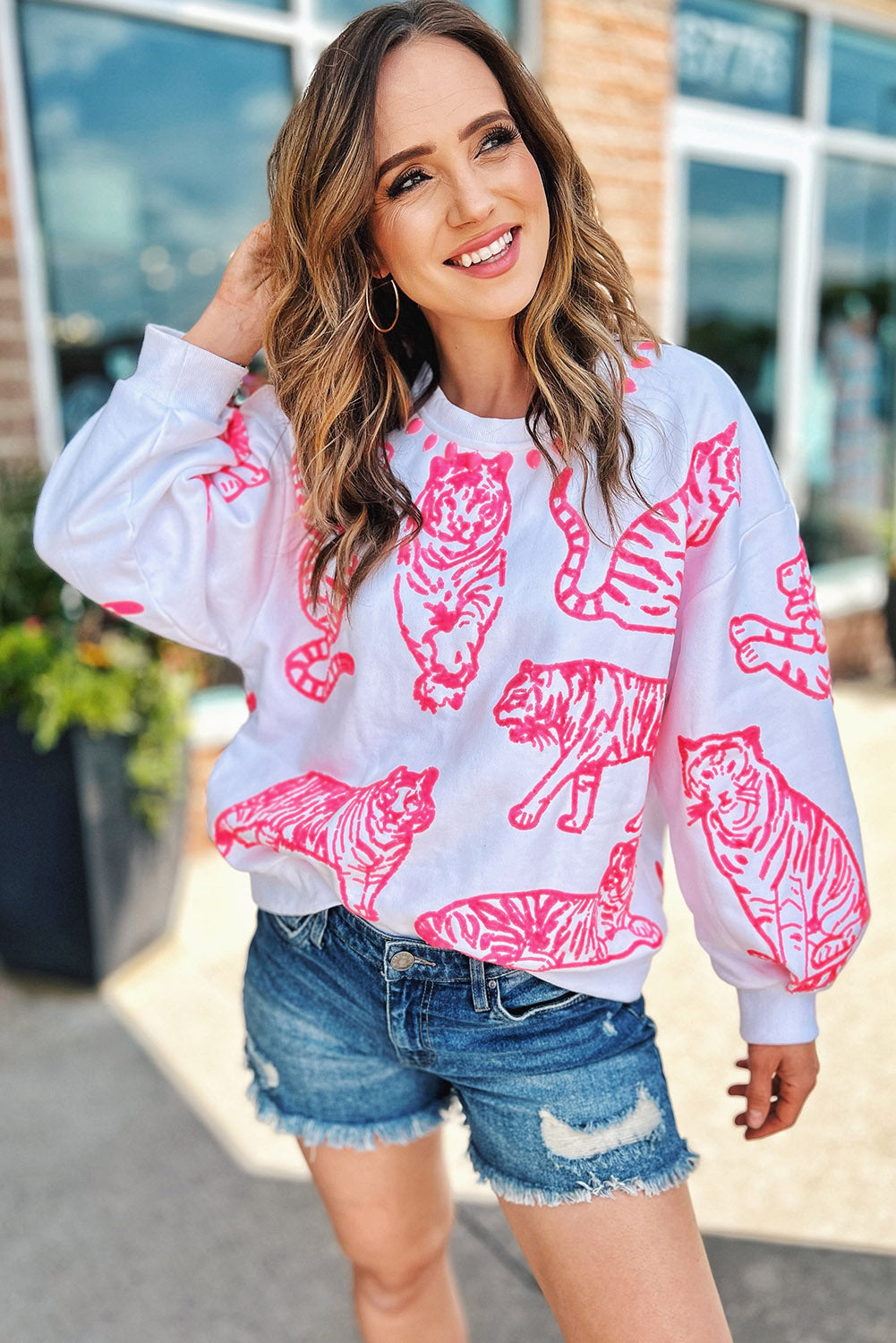 A person with long blonde hair wears a trendy Bright White Pink Animal Print Pullover Sweatshirt, paired with blue jeans, standing outdoors with their back facing the camera.