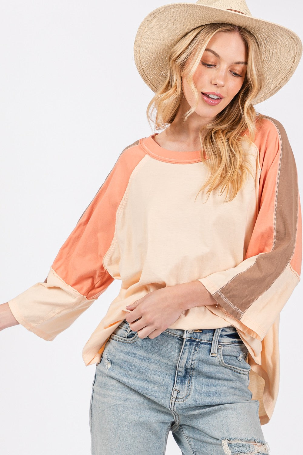 A woman dons a wide-brimmed hat and a modern look with her SAGE + FIG Color Block Curved Hem T-Shirt paired effortlessly with jeans, creating a versatile piece that showcases her vibrant style.