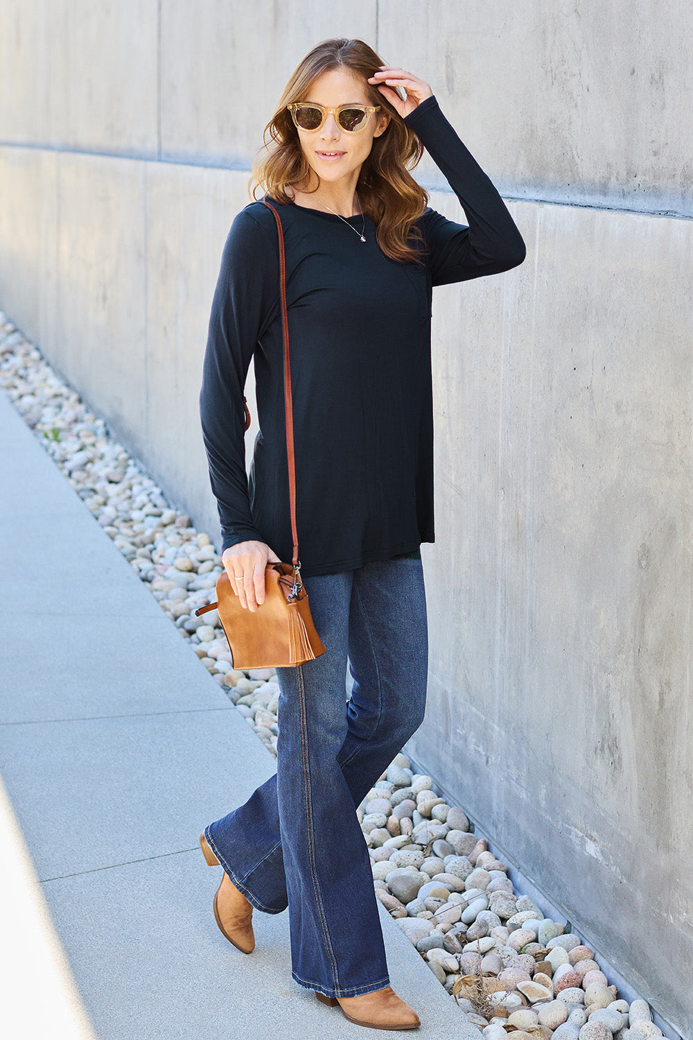 A woman with long hair wearing sunglasses, a Basic Bae Full Size Round Neck Long Sleeve Top in turquoise, and jeans is standing in front of a concrete wall with one hand on her hip.