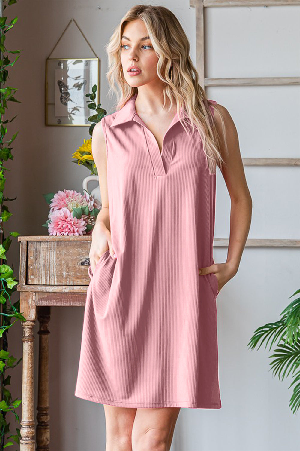 A woman in a versatile Heimish Full Size Ribbed Johnny Collar Sleeveless Dress and straw hat stands by a window, looking outside, with one hand in her pocket.