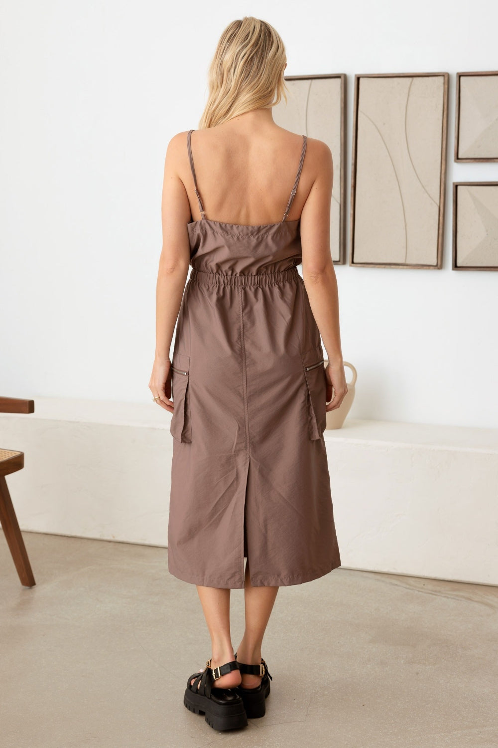 A woman stands indoors wearing the Le Lis Adjustable Strap Waist Drawstring Cargo Midi Dress paired with black platform sandals. She poses with her hands on her hips, showcasing the sleeveless brown dress with a cinched waist.