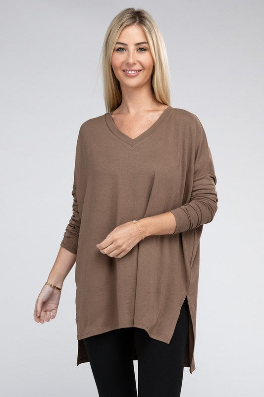 A woman with long, blonde hair is wearing a loose-fitting, brown Dolman Long Sleeve V-Neck Side Slit Hi-Low Hem Top and black pants. She poses with one hand on her hip and a neutral facial expression against a light grey background.