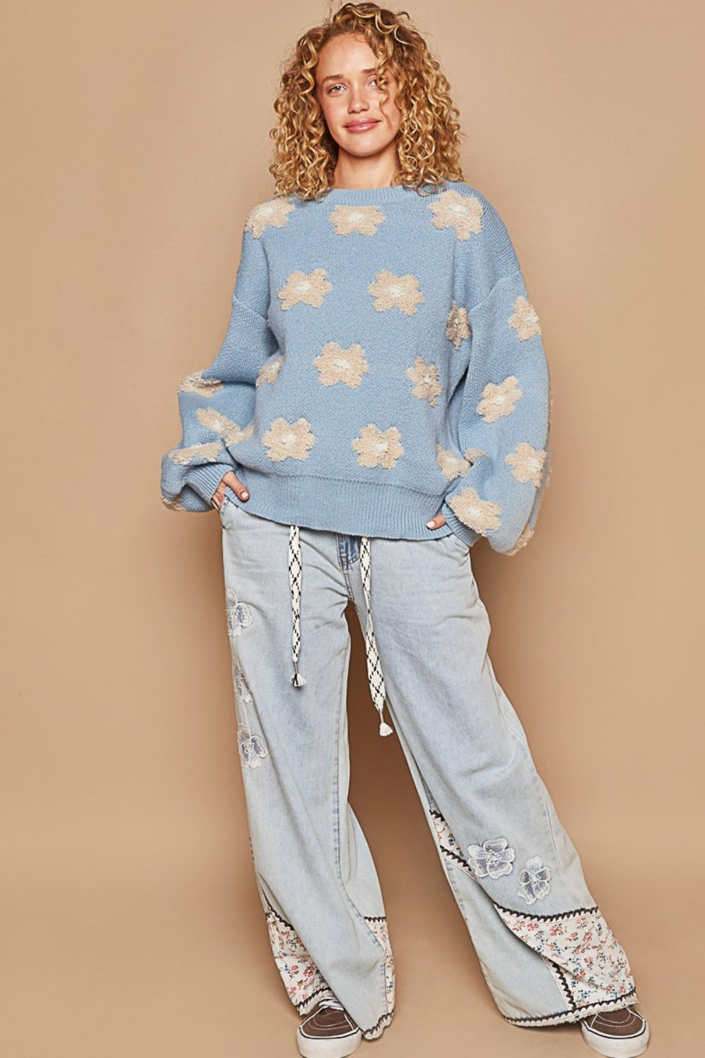 A person with curly hair sits against a matching beige background, wearing the oversized POL Daisy Pattern Drop Shoulder Sweater in blue adorned with beige daisy patterns.