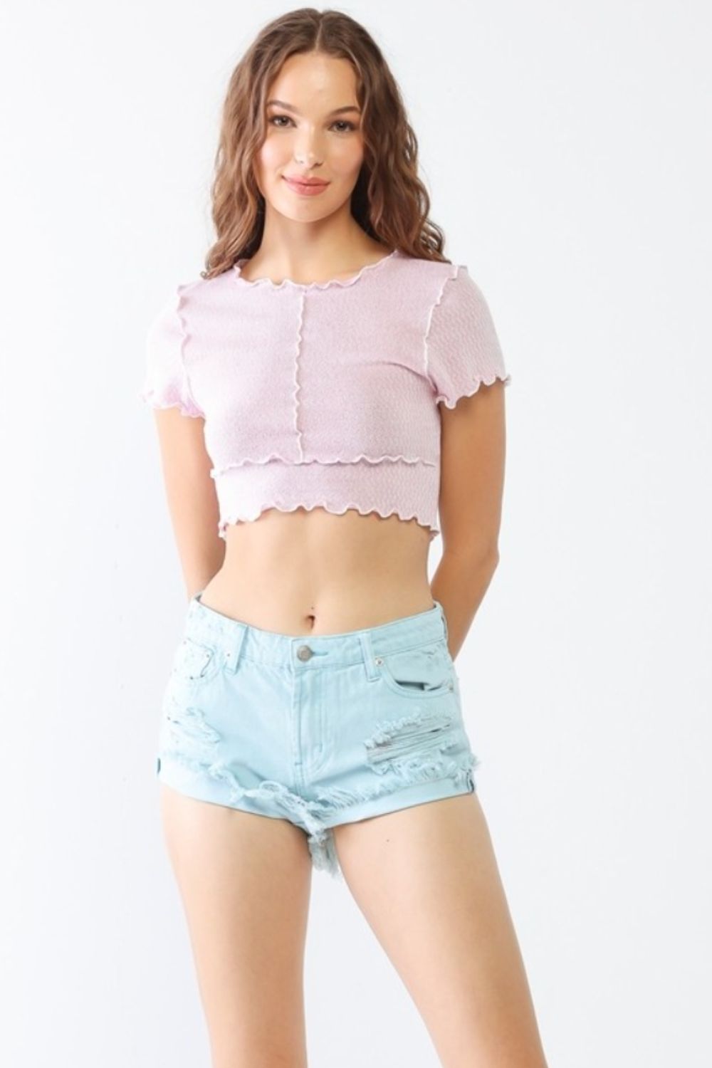 A person stands against a plain background, wearing a light pink crop top and LITZ LA Distressed Ripped Denim Shorts. They have long, wavy hair, embracing a trendy summer style.