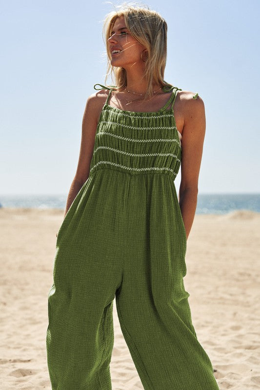 A person wearing the Washed Multi Smocked Detail Tie Straps Jumpsuit stands on the sunny beach, with the ocean creating a perfect backdrop.