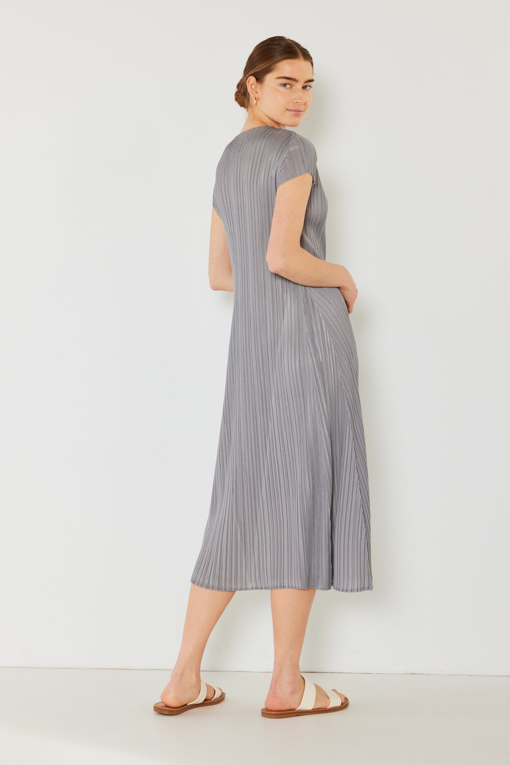 A person stands against a light wall, wearing an elegant Marina West Swim Pleated Cap Sleeve A-Line Dress in black and white sandals.