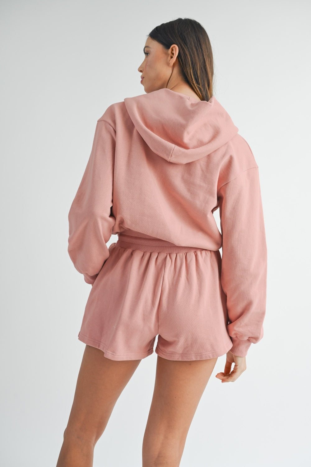 Woman wearing a MABLE French Terry Hooded Romper in pink, hands in pockets, looking directly at the camera.