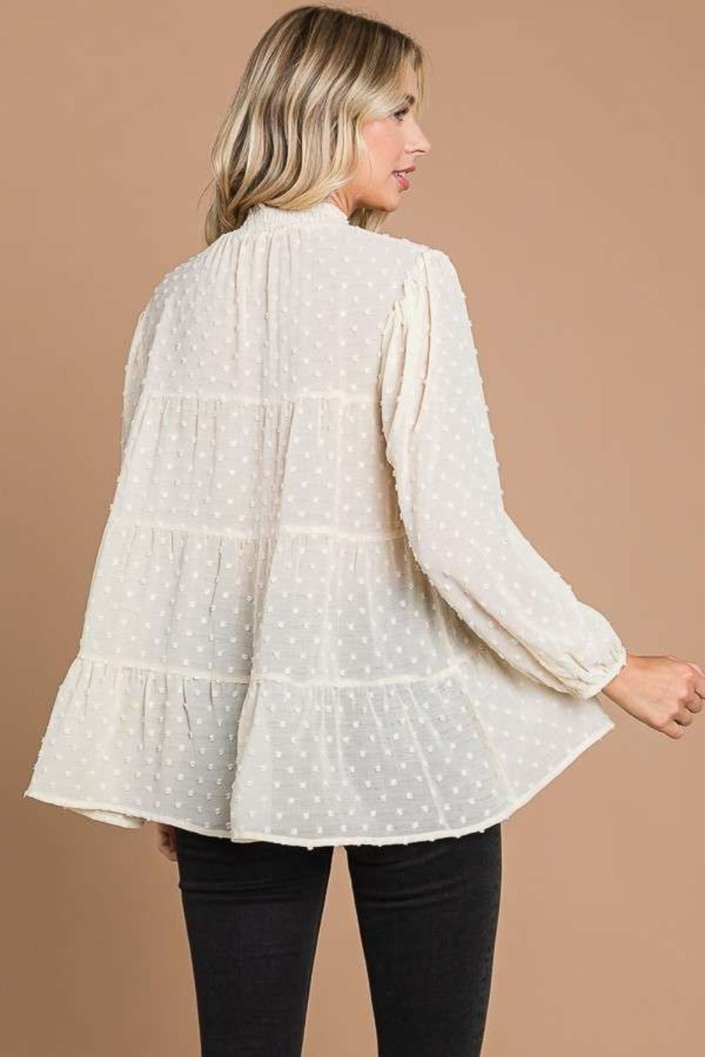 A woman with blonde hair stands against a plain brown background, exuding feminine fashion elegance in a loose, long-sleeved Culture Code Full Size Swiss Dot Smocked Mock Neck Blouse.