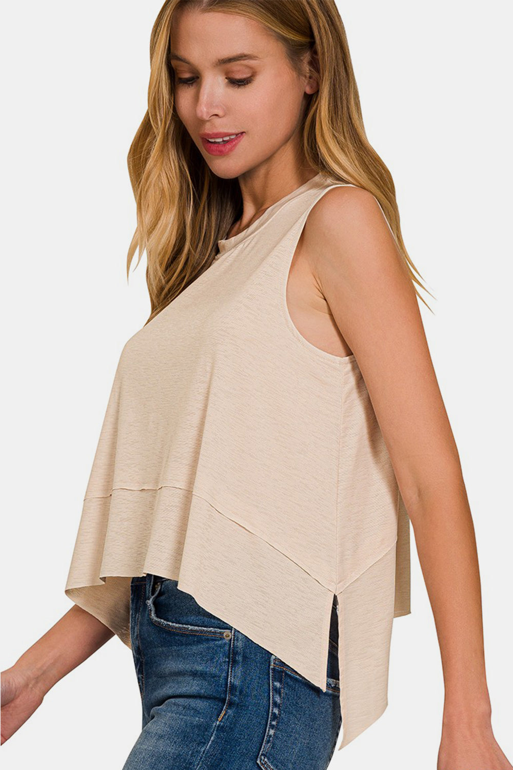 A woman with long, light brown hair wears a versatile piece: the Zenana Exposed Seam Slit Round Neck Tank in beige paired with blue jeans, standing against a plain background.