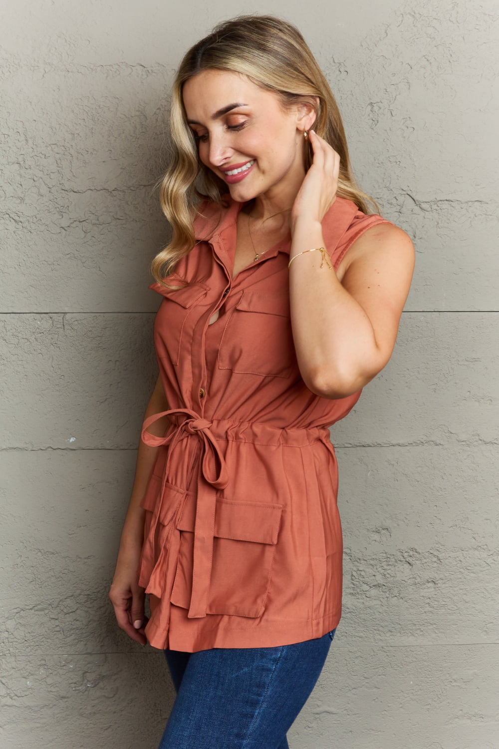 A woman wearing the Ninexis Follow The Light Sleeveless Collared Button Down Top in rust-orange with a tied waist poses in front of a textured grey wall, touching her hair while looking down. She pairs the top with blue jeans featuring an elastic waistband.