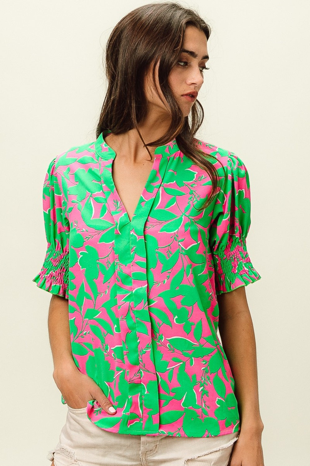 A woman wearing the BiBi Smocked Cuff Floral Contrast Top, featuring a chic design with bright pink and green floral patterns and ruffled sleeves, poses with one hand near her chin.