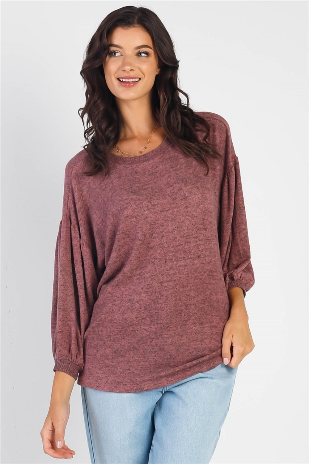 A person with long dark hair stands against a white background, smiling and raising one hand while wearing the trendy and stylish Cherish Apparel Drop Shoulder Puff Sleeve Top in burgundy paired with light blue jeans.