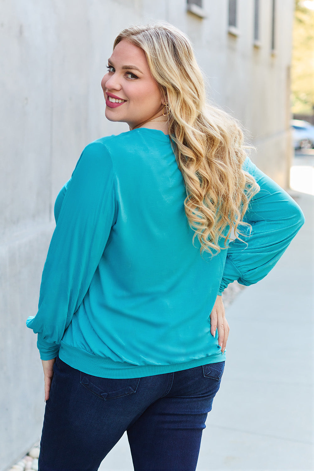 A woman with brown hair is standing against a concrete wall, wearing the Basic Bae Full Size V-Neck Lantern Sleeve Top in teal and blue basic-style jeans, smiling.