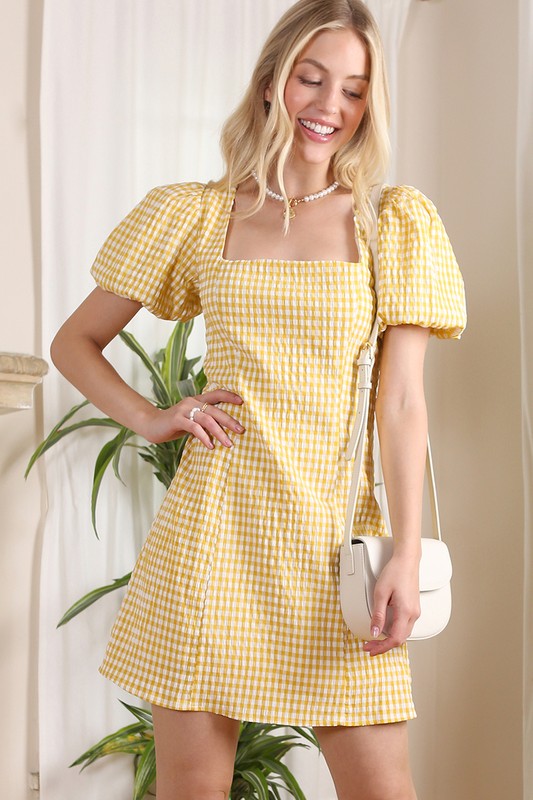 A woman stands indoors, smiling while wearing the SS back strap dress in yellow gingham, and holding a white purse.