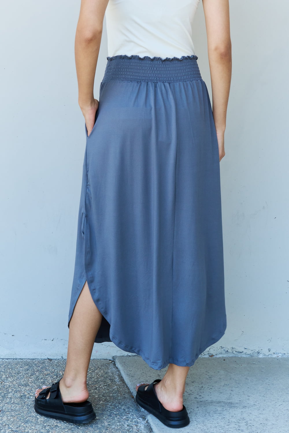 A person wearing a white sleeveless top and the Doublju Comfort Princess Full Size High Waist Scoop Hem Maxi Skirt in Charcoal stands against a plain wall. They are also wearing black sandals.