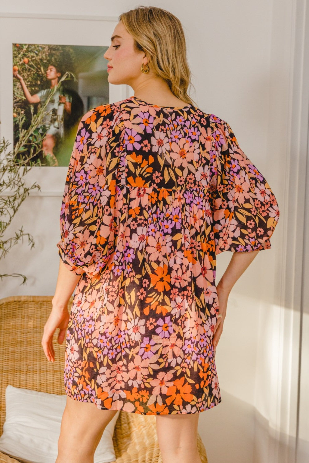 A woman in a flattering silhouette stands indoors with arms folded, wearing the vibrant ODDI Full Size Floral Tied Neck Mini Dress. She poses gracefully in front of a framed picture and wicker chair.