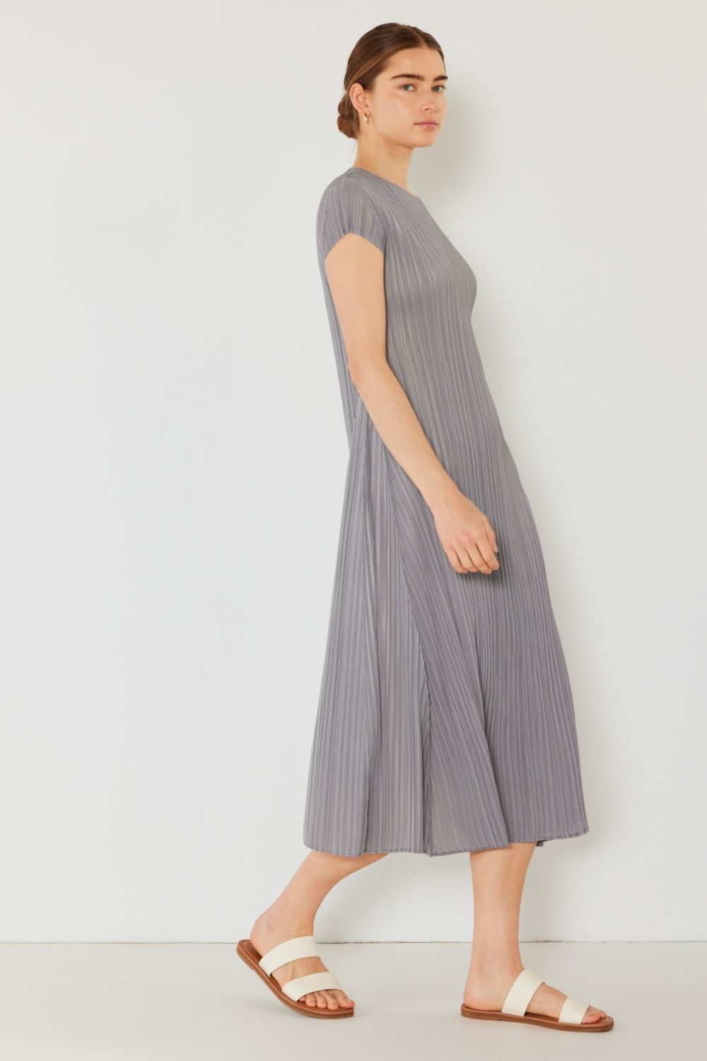 A person stands against a light wall, wearing an elegant Marina West Swim Pleated Cap Sleeve A-Line Dress in black and white sandals.