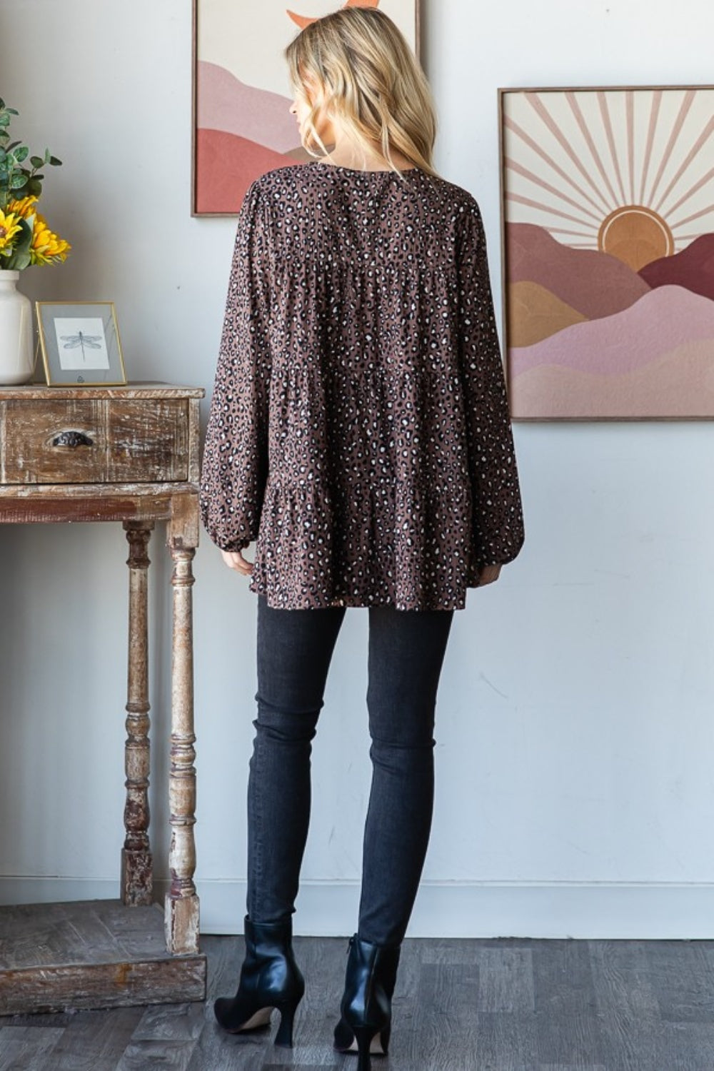 A woman wearing a Heimish Leopard Long Sleeve Tiered Blouse and black pants stands by a window indoors, looking at the camera—embodying a chic and feminine wardrobe.