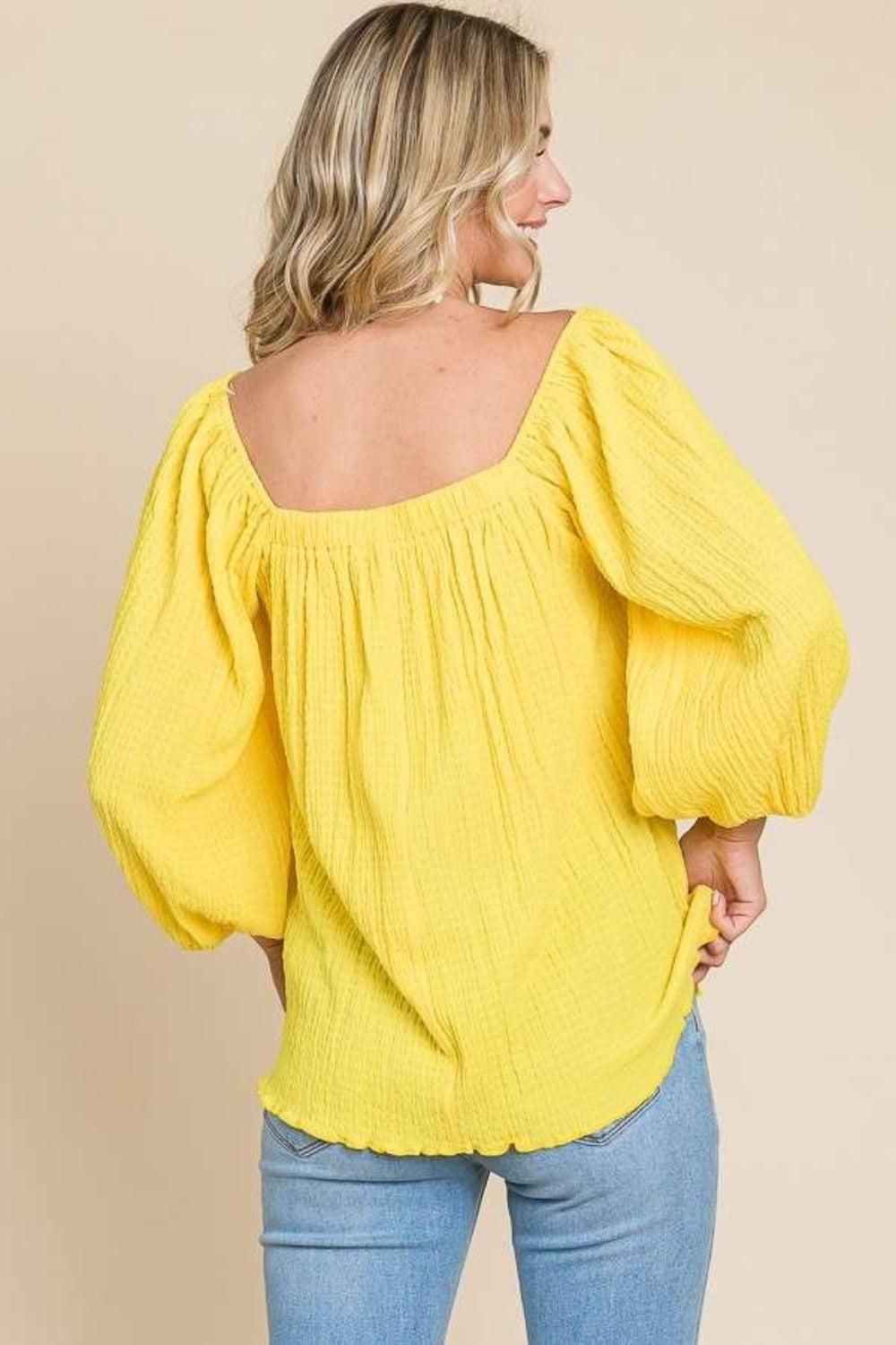 A woman wearing a yellow Culture Code Texture Square Neck Puff Sleeve Top and blue jeans stands against a beige background.