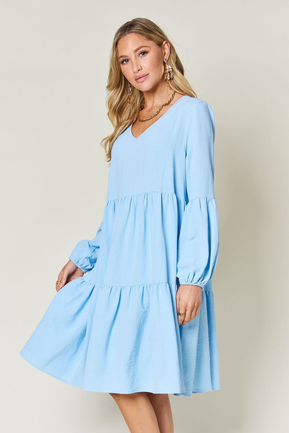 A woman stands wearing the Double Take Full Size V-Neck Balloon Sleeve Tiered Dress with Pockets in pink. Crafted from high stretch yarn, this dress combines comfort and style effortlessly. She has long, wavy hair and accessorizes with hoop earrings and a necklace. This imported garment is easy to care for – simply machine wash cold to keep it looking fresh.