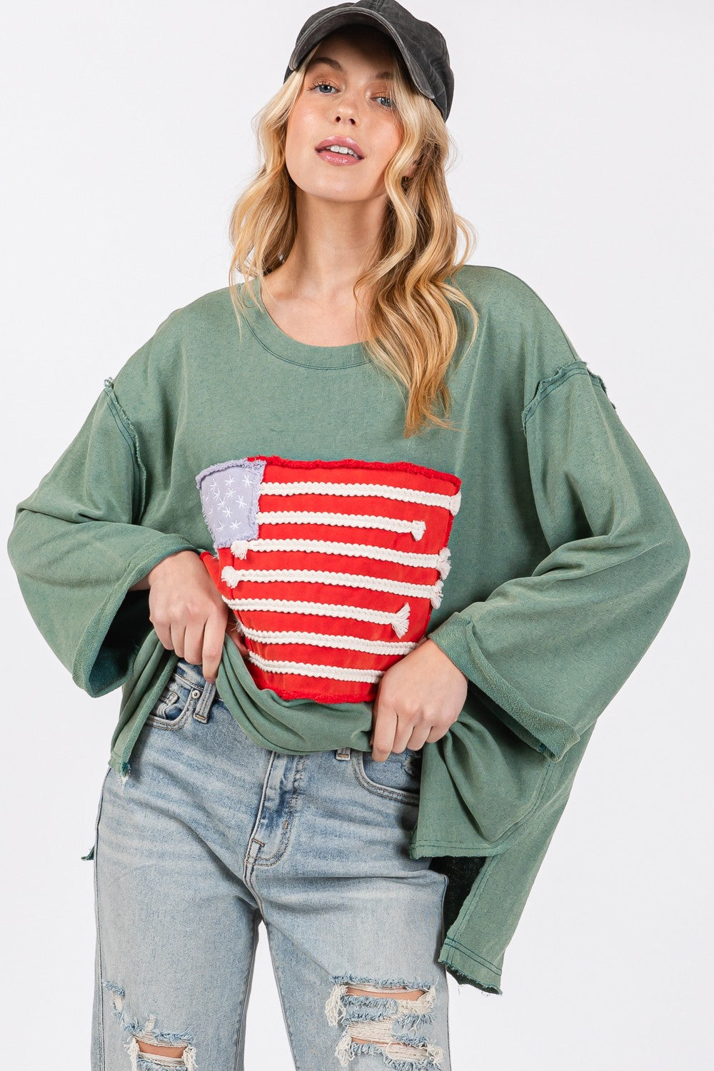 A person exudes casual charm in the SAGE + FIG Full Size American Flag Patch Drop Shoulder T-Shirt, paired with ripped jeans, confidently posing with hands in hair against a plain background.