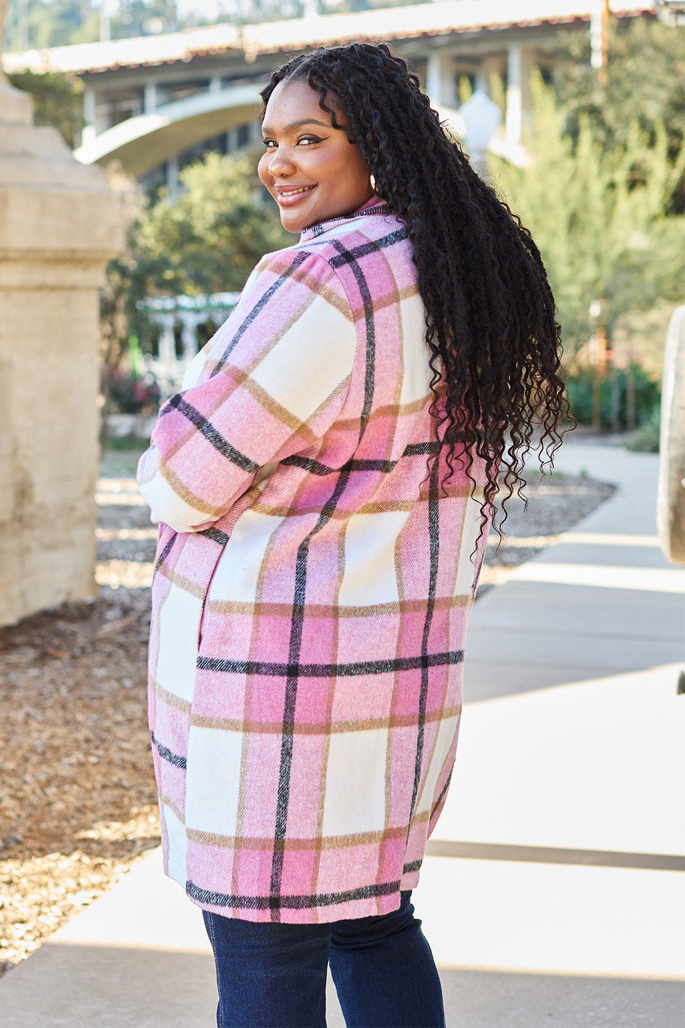 A woman stands on a sidewalk wearing the Double Take Full Size Plaid Button Up Lapel Collar Coat in pink and white over a white top and blue jeans, with her right hand lightly touching her hair. The polyester fabric of the coat makes it durable and perfect for an easy machine wash cold.