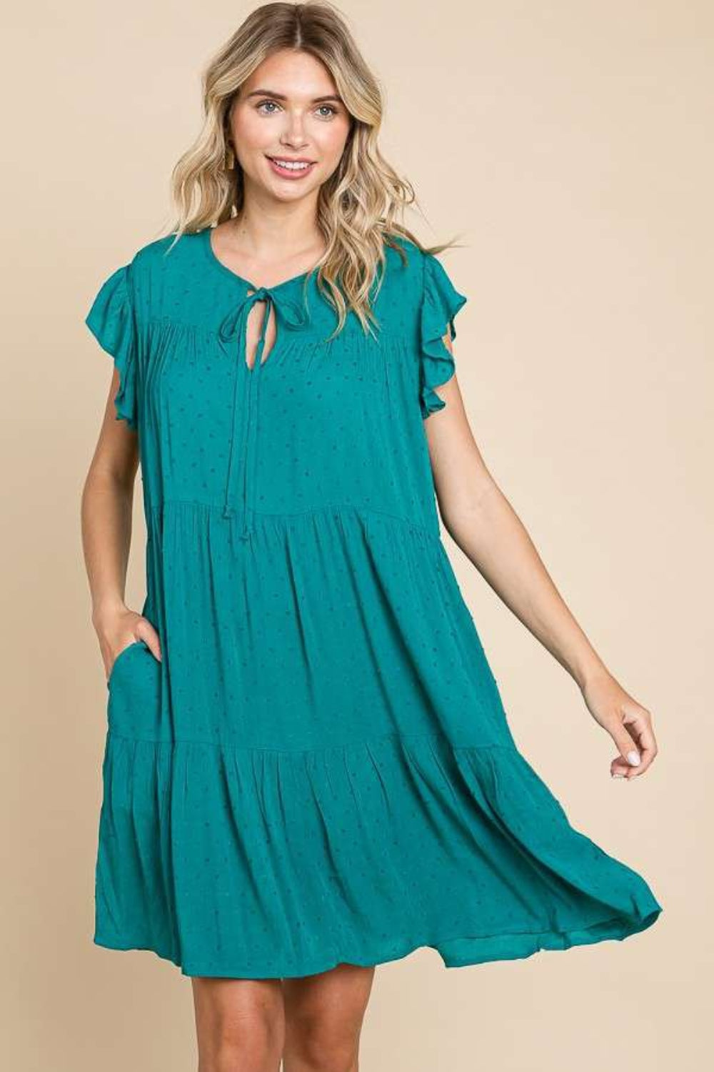 A woman stands against a beige background wearing the Culture Code Ruffle Cap Sleeve Tiered Dress, which is teal and flowing with short ruffled sleeves and a tie at the neckline. She is smiling with one hand in her pocket, showcasing the lightweight fabric.