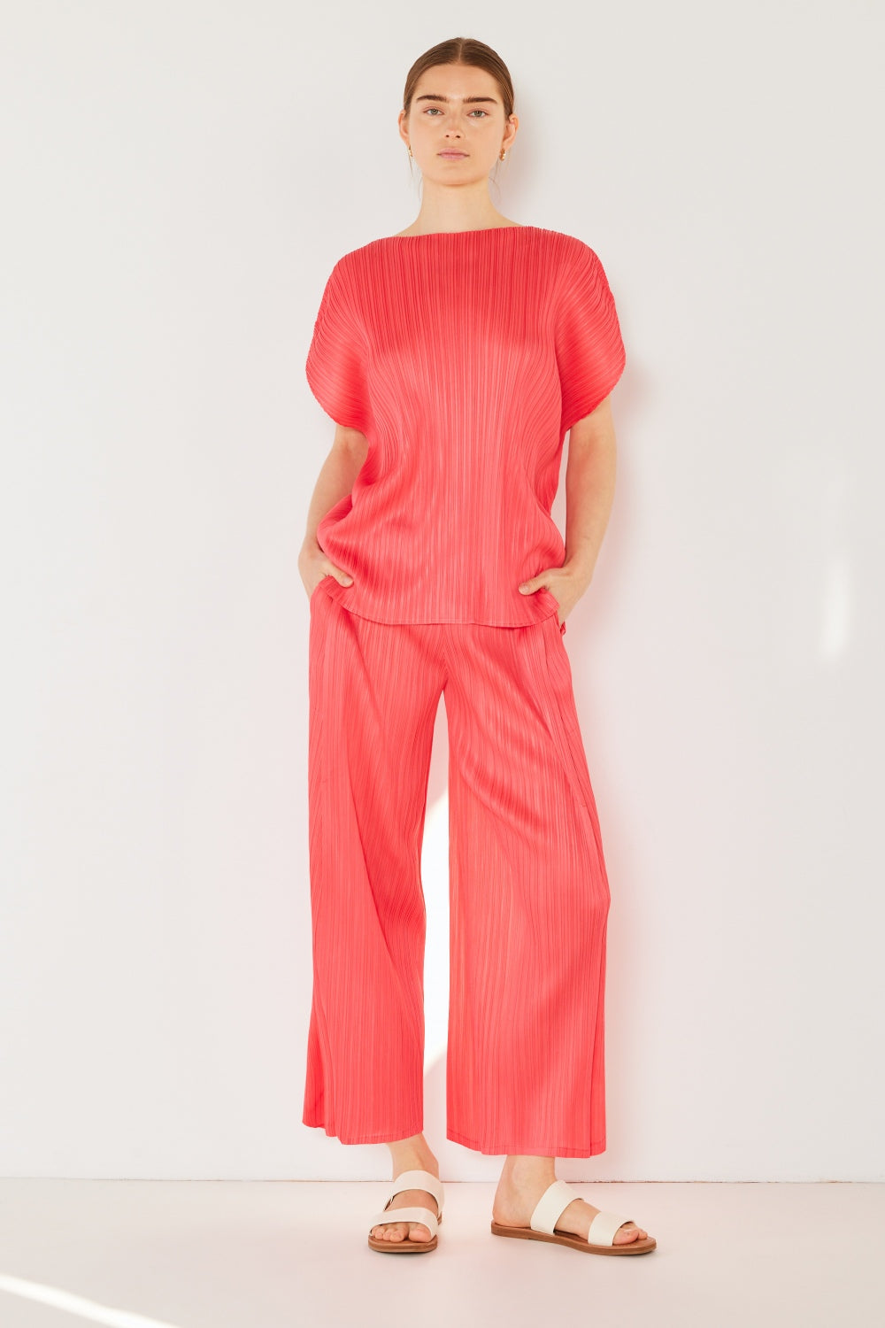 A person wearing the Marina West Swim Pleated Wide-Leg Pants with side pleat detail in red, standing against a white background with hands in pockets and wearing white sandals.