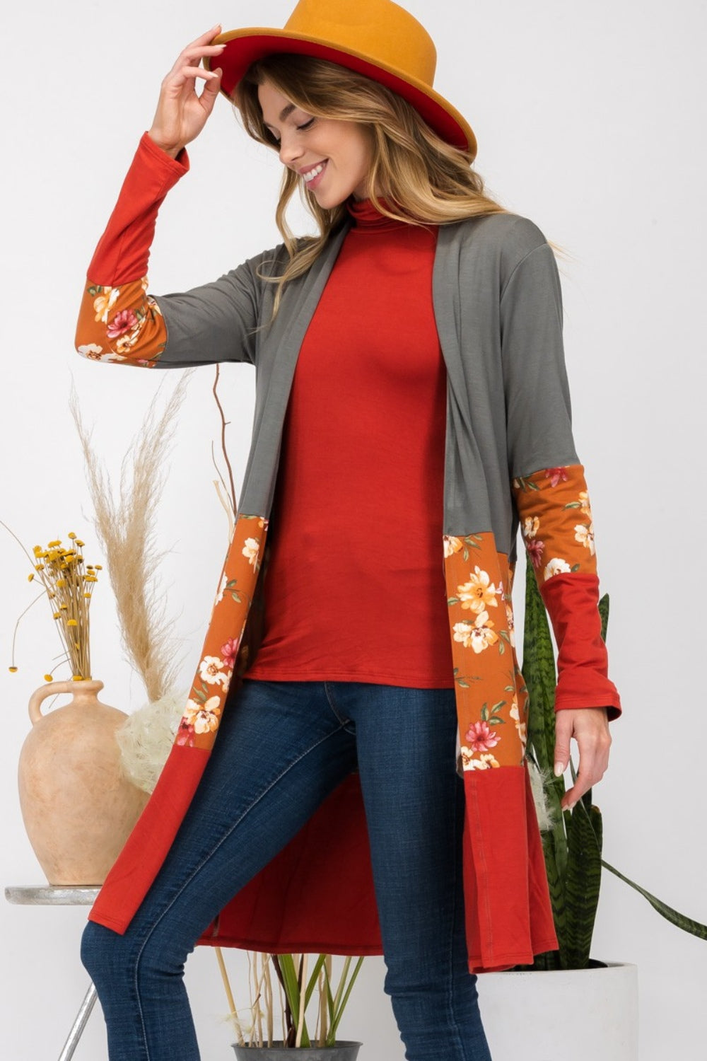 Wearing the Celeste Full Size Floral Color Block Open Front Cardigan, a person in a red hat stands indoors near potted plants, smiling.