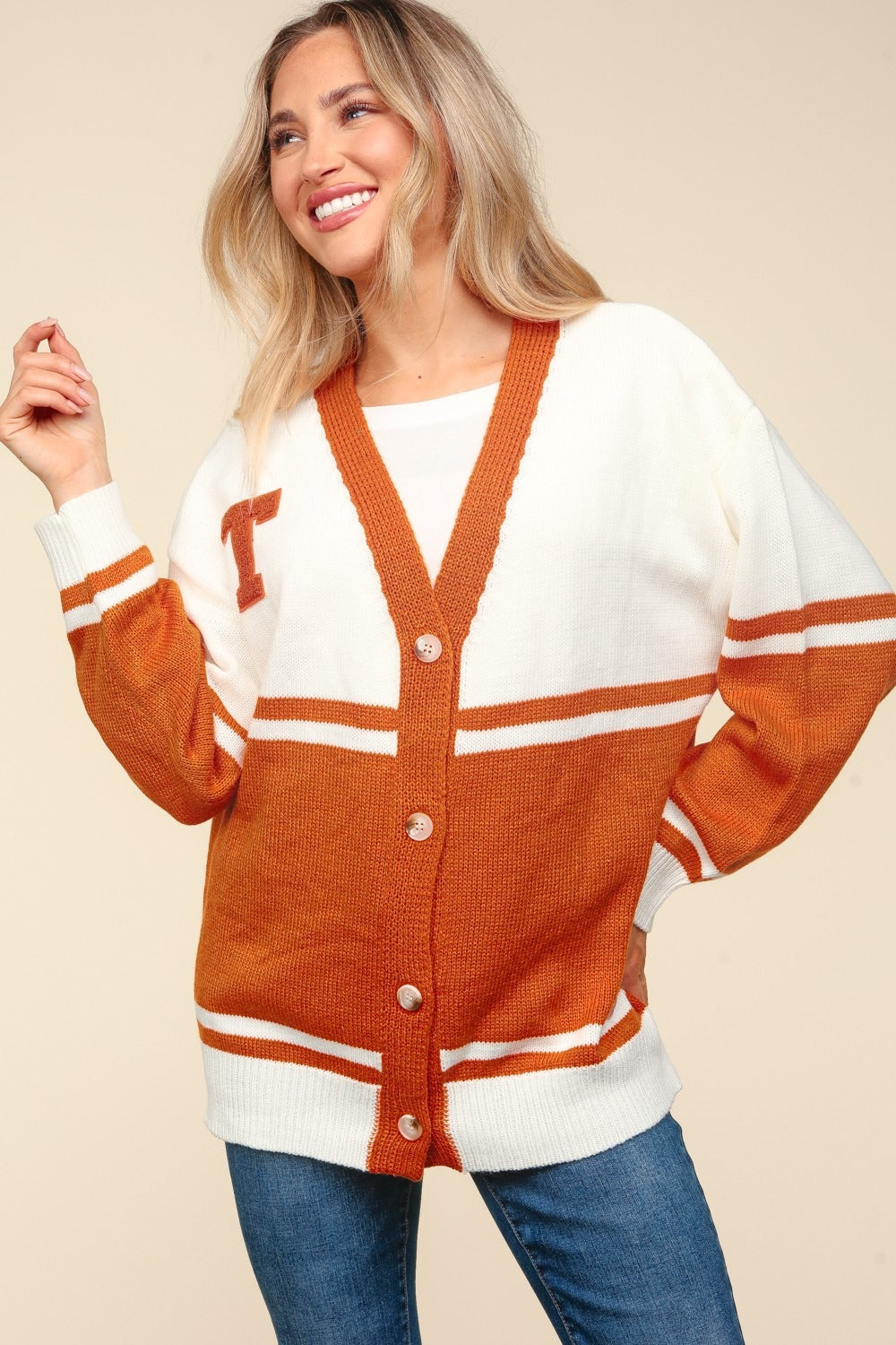 A smiling woman wearing the Haptics Full Size V Neck Button Down Letter Patch Cardigan, an oversized fit in white and orange with a "T" patch, poses with one hand on her hip and the other touching her hair.