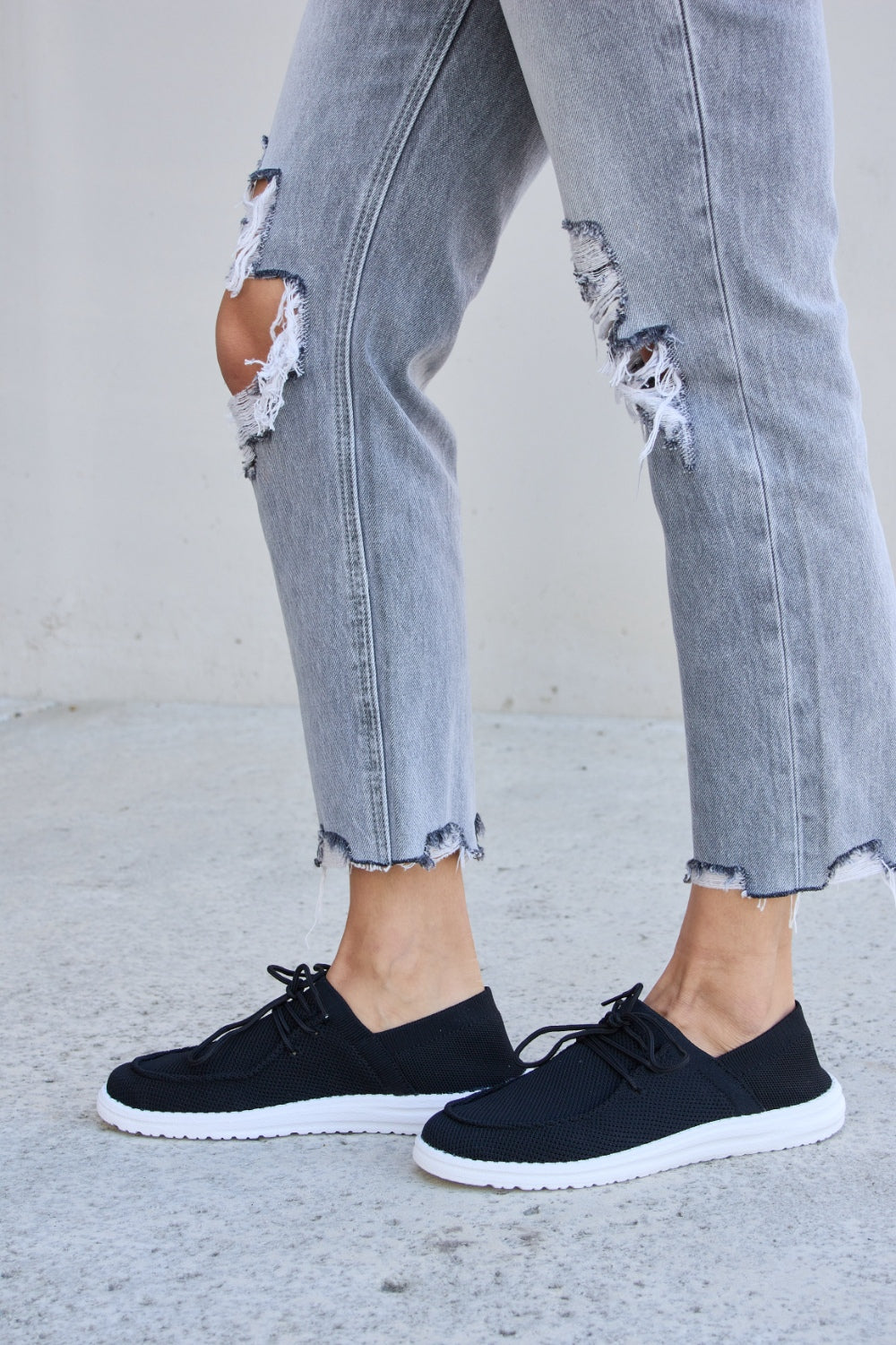 A person wearing ripped jeans and Forever Link Flat Round Toe Lace-Up Sneakers with white soles stands on a concrete surface.