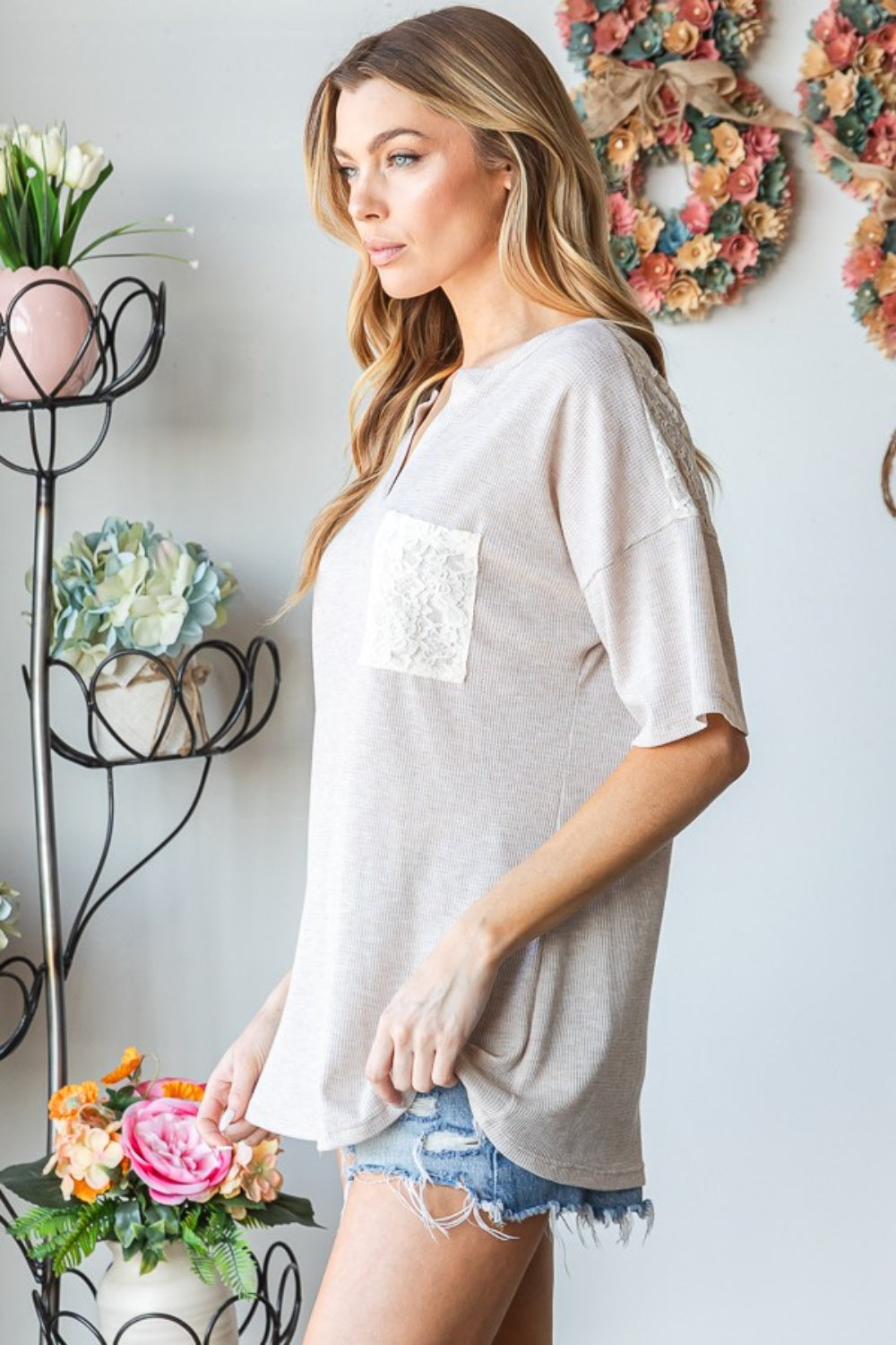 A woman wearing the Heimish Full Size Lace Front Pocket Drop Shoulder Top stands indoors near a decorative metal stand adorned with flowers.