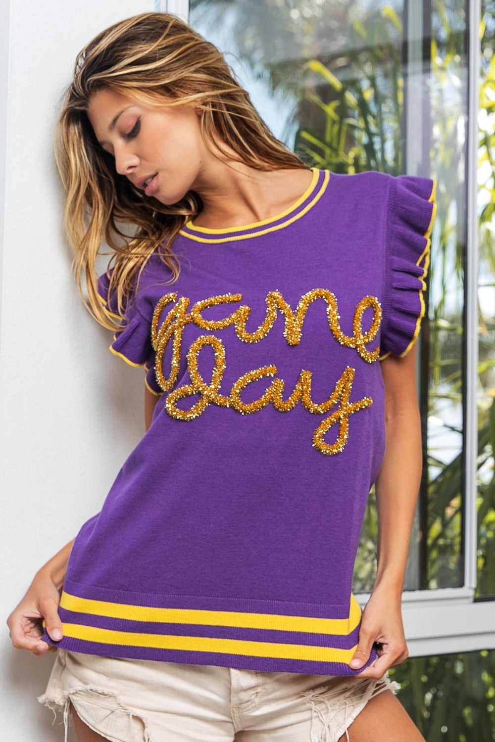 A person wearing the BiBi Game Day Letter Contrast Trim Ruffled Sleeveless Sweater, with its purple color and gold "Game Day" lettering, gestures towards the text. They stand by a window, pairing it with white shorts that accentuate their lively ensemble's contrast trim.