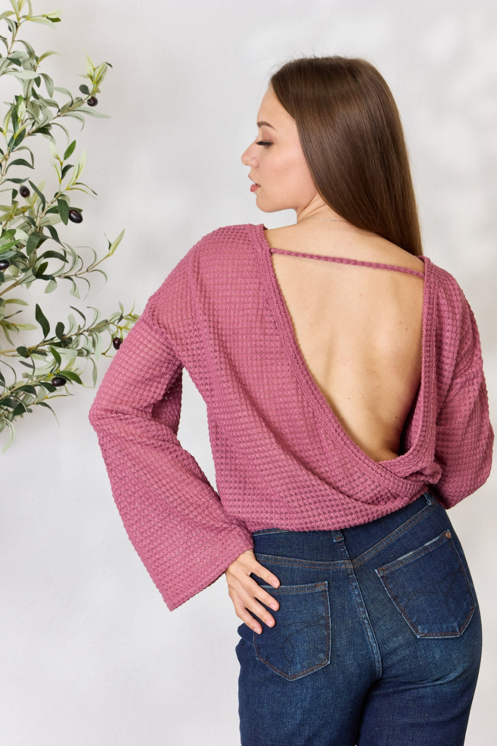 A person wearing a Culture Code Full Size Waffle-Knit Round Neck Long Sleeve Blouse in pink and jeans stands in front of a light background, complemented by a leafy plant on the left.