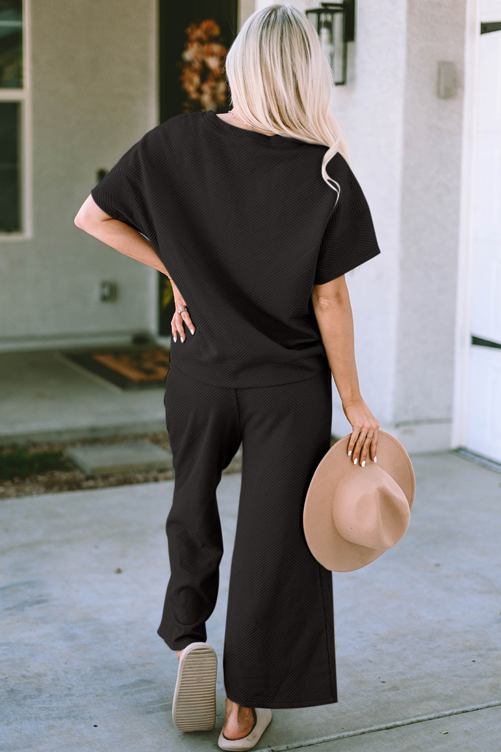 A woman with long hair smiles while standing outside a building, dressed in the Double Take Full Size Texture Short Sleeve Top and Pants Set, featuring a matching beige two-piece ensemble. She wears slides and holds a hat in her right hand. Her outfit, slightly stretchy for added comfort, is perfect for machine wash cold.